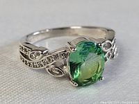 Front and angled views of the sterling silver ring with an oval green stone and clear accent stones on a detailed band.