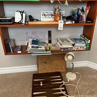 View of shelf with assorted office items including pens, pencils, books, greeting cards, calculators, binoculars, and other supplies. Below shelf is backgammon set and white desk lamp.