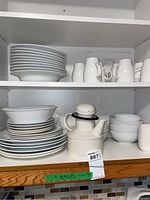 White plates, bowls, mugs, and teapot stacked in a cabinet shelf. Visible variety of dish types and shapes.