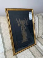 Full view of framed brass etching on a couch showing gold frame and dark matte background.