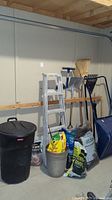 Photo showing assorted gardening supplies and tools against a wall: including aluminum ladder, brooms, rakes, garden tools, bags of soil and mulch, planters, black trash bin.