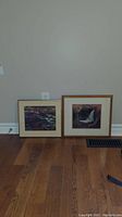 Two framed prints leaning against wall on hardwood floor showing two landscape art pieces.