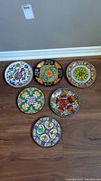 Six hand-painted ceramic plates arranged on wood floor showcasing colorful floral and fruit designs from Mexico, Spain, and Greece.