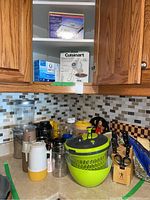 Kitchen countertop with various kitchenware items including a salad spinner, plastic bowls, thermoses, brass board, canisters, storage containers, and a knife block with knives visually identifiable.