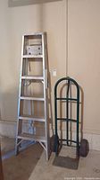 Front view of 6-foot aluminum A-frame ladder standing upright next to a green metal hand truck with two large black tires.