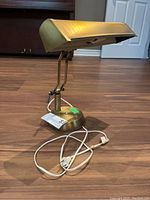 Front and side view of a brass banker’s lamp with green glass shade on wooden floor, showing electrical cord and adjustment lever.