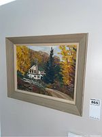 Framed painting hanging on a wall showing a white house surrounded by autumn foliage and a forested pathway.