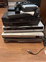 Stacked electronics including Toshiba VCR player, Panasonic DVD recording machine, Panasonic DVD CD player, Motorola HDTV receiver, Samsung DVD player, with remote controls on top.