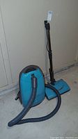 Full view of teal Kenmore vacuum cleaner with hose and blue wide floor attachment standing upright on concrete floor.