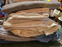 Stack of folded bed linens including flat and fitted sheets with pillowcases visible, primarily neutral colors including beige and gray.