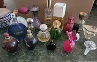 Overview of multiple fragrance bottles on a surface, showing varied shapes, colors and conditions of bottles, some with spray tops and two loose stoppers.