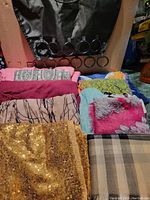 Several folded scarves and pashminas displayed in varying colors and patterns including gold sequins, plaid, and floral prints with a black Umbra scarf hanger with circular rings in the background.