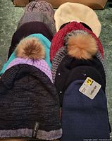 Multiple ladies' winter hats stacked together showing different colors, designs, and some with faux fur pom-poms.