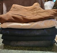 Stack of five pairs of men's pants in varying colors and textures.