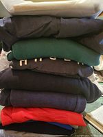 Stack of folded men's clothing including jackets, shirts, track pants in dark colors mostly with one red garment at bottom.