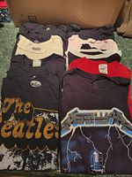 Lot of folded assorted musical artist t-shirts in dark and light colors including Beatles and Metallica, laid on a dark surface.