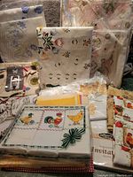 Photo showing assorted linens including aprons, tablecloths with floral and abstract prints, and napkins arranged together on a surface.