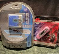 Sony Walkman MiniDisc player and Nexstar MP3 player in original sealed packaging placed side by side on a green surface.