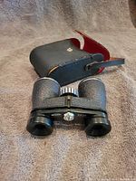 Binoculars sitting in front of an open black case with a red interior on a beige background.