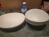Two large white serving bowls on left, stacked four white cereal or salad bowls on right, water bottle behind for scale