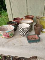 Photo showing a set of 10 different flower pots made from ceramic, metal, and plastic arranged on a concrete surface outdoors. Variety of floral, geometric, solid color, and plain designs.