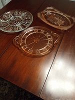 Three pink depression glass serving plates and dishes on wooden table: round divided plate, square dish, and rectangular handled plate