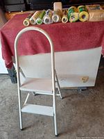 White solid metal two-step ladder with non-slip steps and curved top handle on a concrete floor with red cloth background.