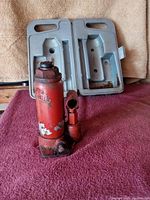Photo showing the red hydraulic bottle jack placed next to the gray metal storage case, highlighting the overall size, color, and condition of the jack and case.