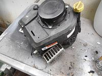 Side view of old black gas lawnmower engine showing recoil starter, yellow fuel cap, and air intake grille in a dirtied condition.