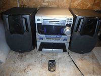 Front view of JVC mini stereo central unit with two speakers and remote control on tiled floor