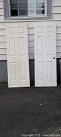 Pair of two 6-panel primed slab doors standing against exterior wall, one used and one new variety visible side by side.