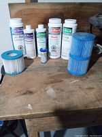 Photo of 5 plastic bottles of spa chemicals including pH Plus, Defoamer, Shock, Filter Cleaner, and test strips alongside three new spa filters on a wooden surface.