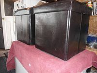 Two black faux leather collapsible stools with lids shown side by side on a pink surface under bright lighting. One stool appears slightly taller than the other.