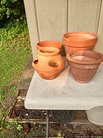 Five terra cotta pots arranged on a table outside; includes decorative pot with handles and standard round pots.