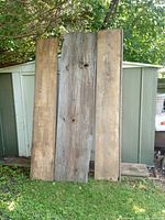 Three wide vintage lumber pieces leaning against a metal shed, two smoother church pew boards and one rougher barnboard center board