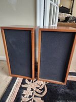 Two KLH Model 06 speakers side by side on carpeted floor against wall showing wooden cabinets and black grilles.