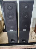 Photo showing a pair of black Sony floor standing speakers with fabric grills and bass ports, standing on carpeted floor.