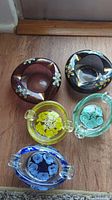 Top-down view of five art glass ashtrays on wooden surface showing varying colors and flower designs