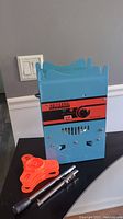 Photo of blue and red bubble tea sealing machine standing upright on black surface with orange plastic base and metal wand accessories next to it.