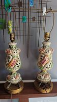 Pair of ornate ceramic table lamps with carved and hand-painted bodies featuring birds and human figures. One lamp has replacement cord and functioning light bulb fitting with missing knob, the other lamp needs plug repair.