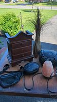 Photo showing wooden jewelry box missing a knob, black leather fanny pack, black cosmetic purse, striped ceramic vase with tall artificial grass, Memorex CD player, and pink salt lamp on a table outside.