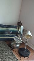 Photo showing three lamps including an antique Coleman Quick-Lite kerosene lamp with metal base, a modern lamp with magnifying glass, and a lamp with white lampshade. Framed artwork is visible behind.