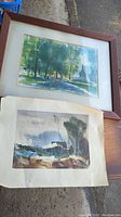 Framed artwork showing outdoor greenery and path, alongside unframed signed print with muted blues and greens
