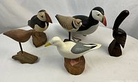 Front and angled view of five wooden bird carvings including Puffins, Seagull, Killdeer, and Sandpiper, showing detailed hand-painting and stands.