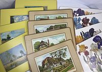 Three sets of dining placemats stacked showing various designs: house and landscape scenes with beige and yellow borders, and floral designs.
