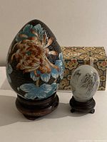 Large cloisonné egg and smaller reverse glass hand-painted egg on wooden stands with patterned box in background
