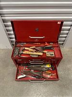 Opened red metal toolbox with several wooden-handled chisels, trowels, screwdrivers and a key on top tray and upper compartment.