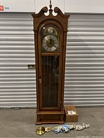 Full view of wooden Bulova grandfather clock with glass front panel, showing the clock case and parts on the floor in front of it.