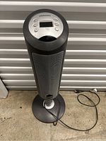 Full view of a black ceramic element tower fan heater standing on the ground. Shows control buttons and LCD screen on top and power cable coiled on floor.