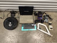 Full lot overview including Toshiba laptop, iRobot Roomba, Logitech keyboard in box, controllers, and other electronics, all placed on cement floor in front of metal door.